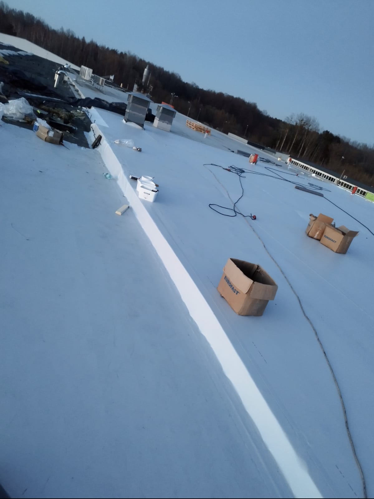 Biała, płaska powierzchnia dachu z detalami montażowymi i kartonami Eurofast, widziana z góry pod kątem, z elementami krajobrazu w tle.