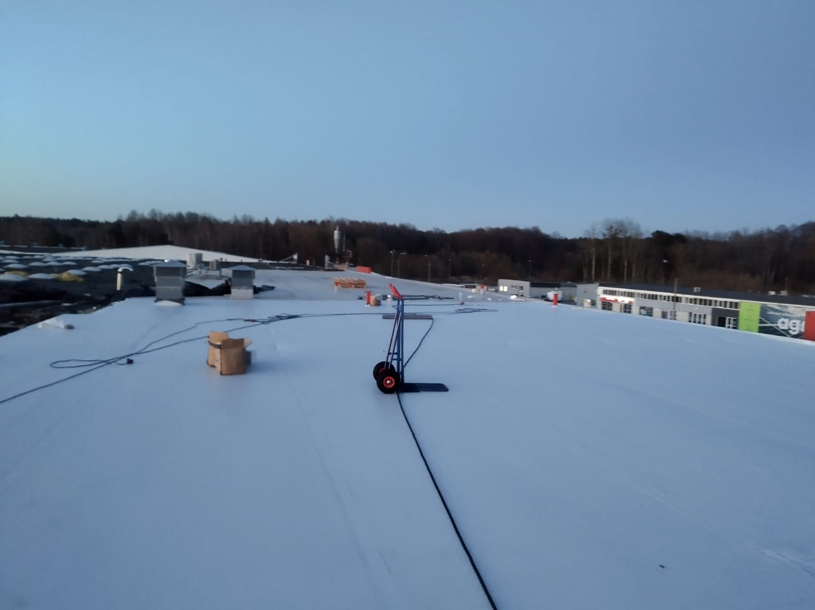 Biała membrana na dachu budynku przemysłowego z wózkami i skrzynką, widok z góry, w tle drzewa i budynki, oświetlenie dzienne.