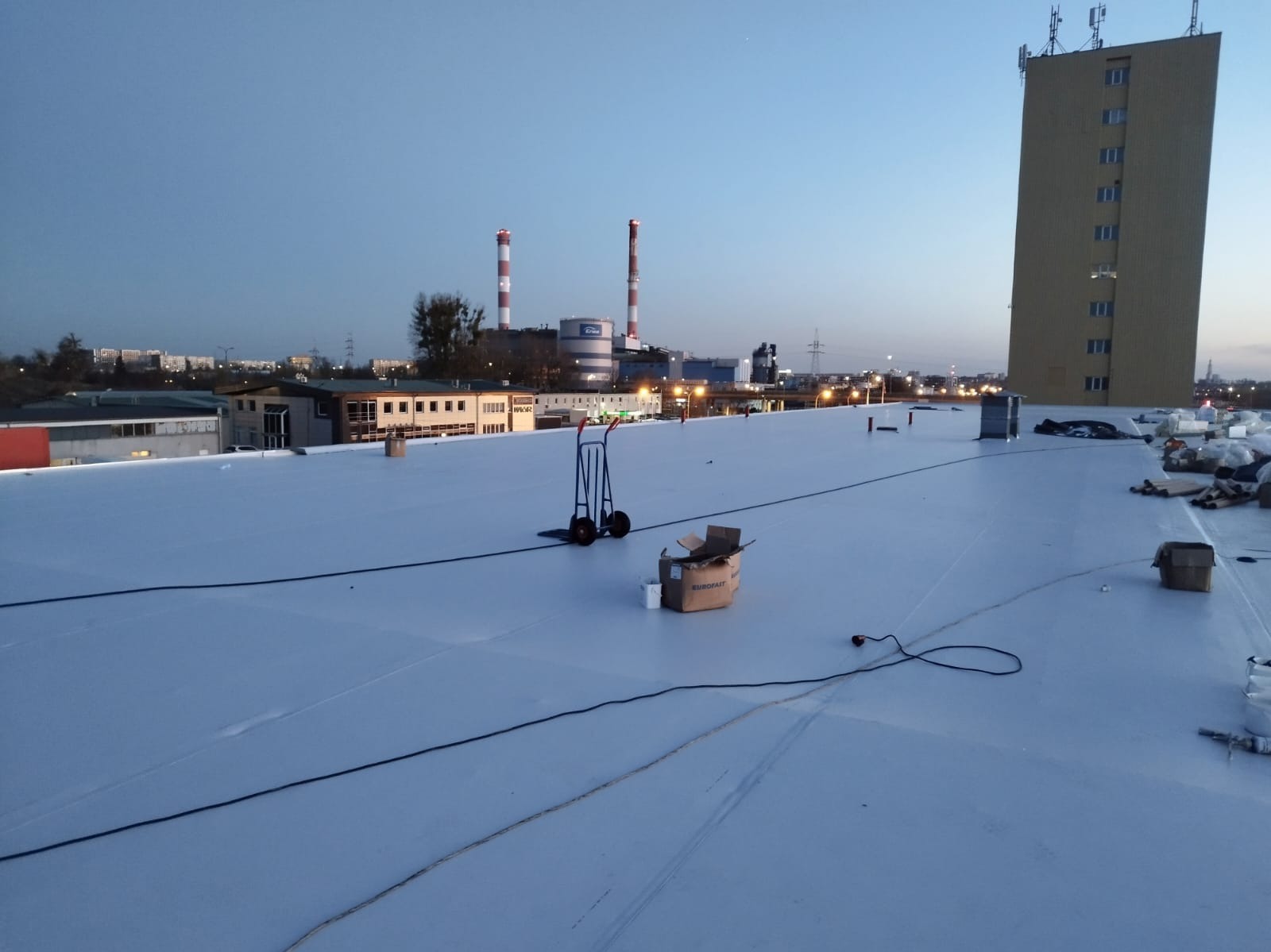 Biała, płaska powierzchnia dachu przemysłowego z widocznymi elementami montażowymi, w tle budynki i kominy. Na dachu wózek transportowy i karton z materiałami.