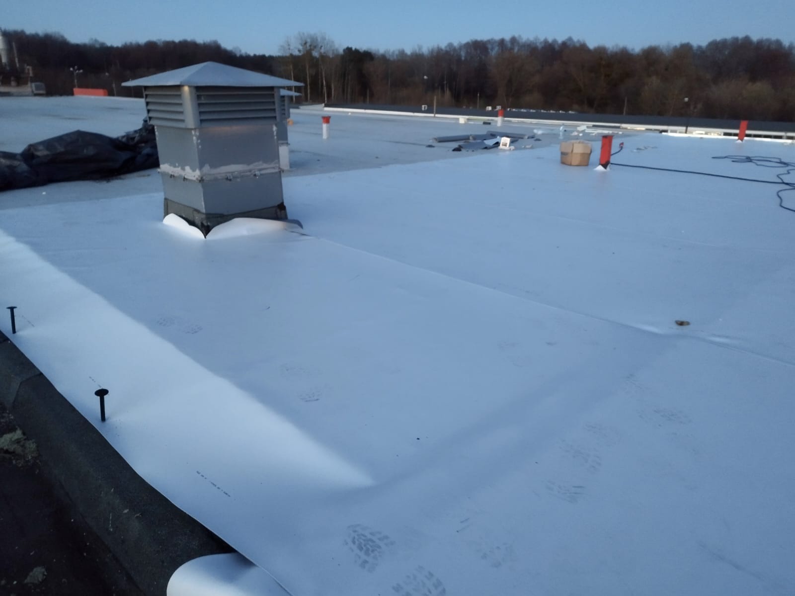 Biała membrana dachowa na płaskim dachu z widocznym kominem wentylacyjnym i śladami stóp. W tle drzewa i elementy instalacji dachowej.