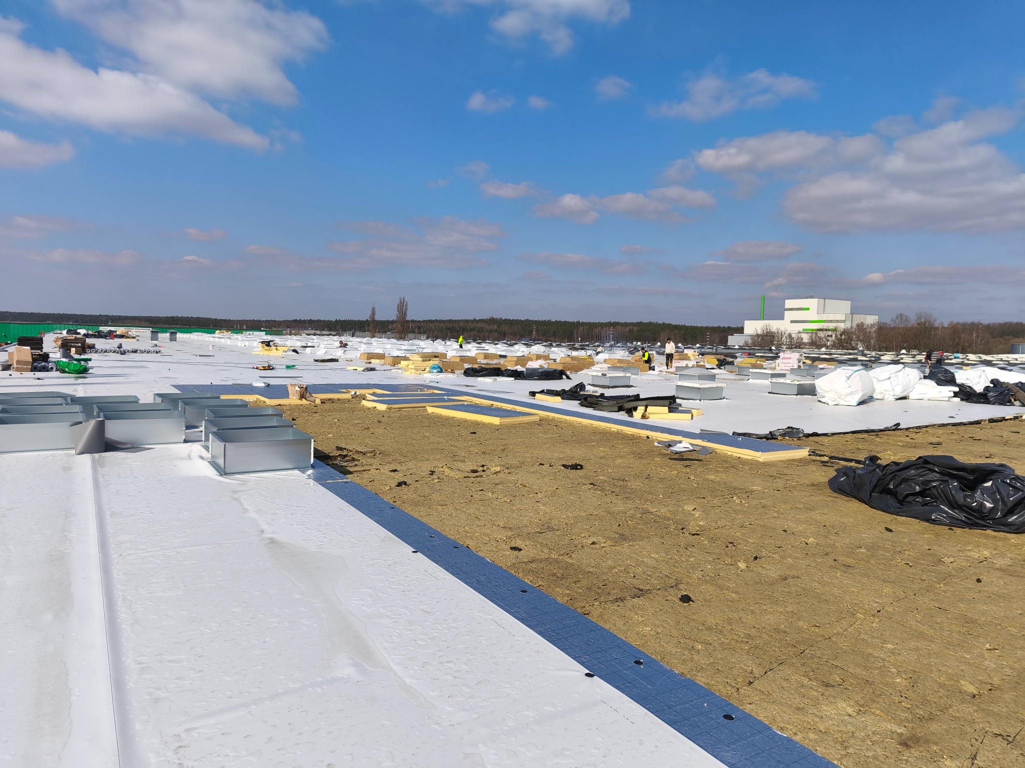 Rozległy dach w trakcie prac: biała membrana, izolacja, elementy wentylacyjne. W tle budynek przemysłowy i błękitne niebo z chmurami. Widoczni pracownicy w odblaskowych kamizelkach.
