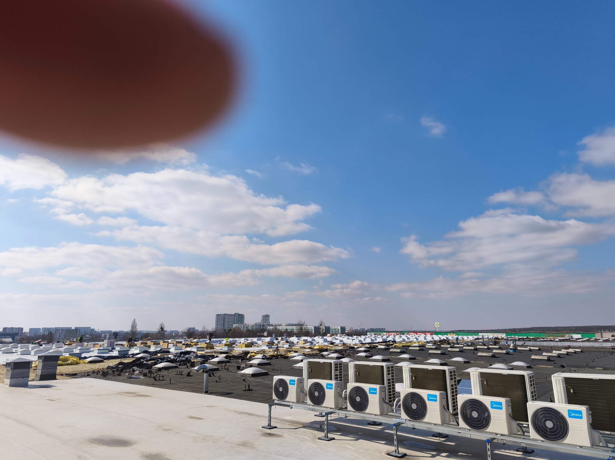 Montaż klimatyzacji na dachu budynku. Widoczne liczne klimatyzatory Midea, elementy konstrukcyjne dachu, miasto w tle i błękitne niebo z chmurami.