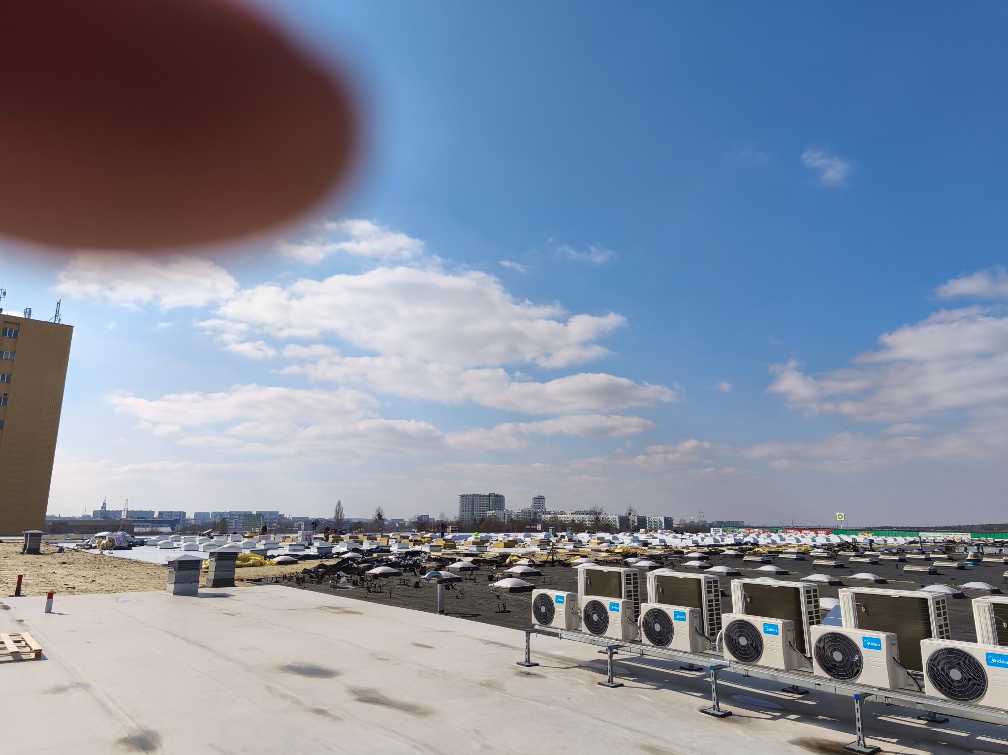 Rozległy dach płaski z licznymi klimatyzatorami Midea w rzędach, w tle widok na miasto pod błękitnym niebem z chmurami. Elementy konstrukcyjne dachu w trakcie montażu.