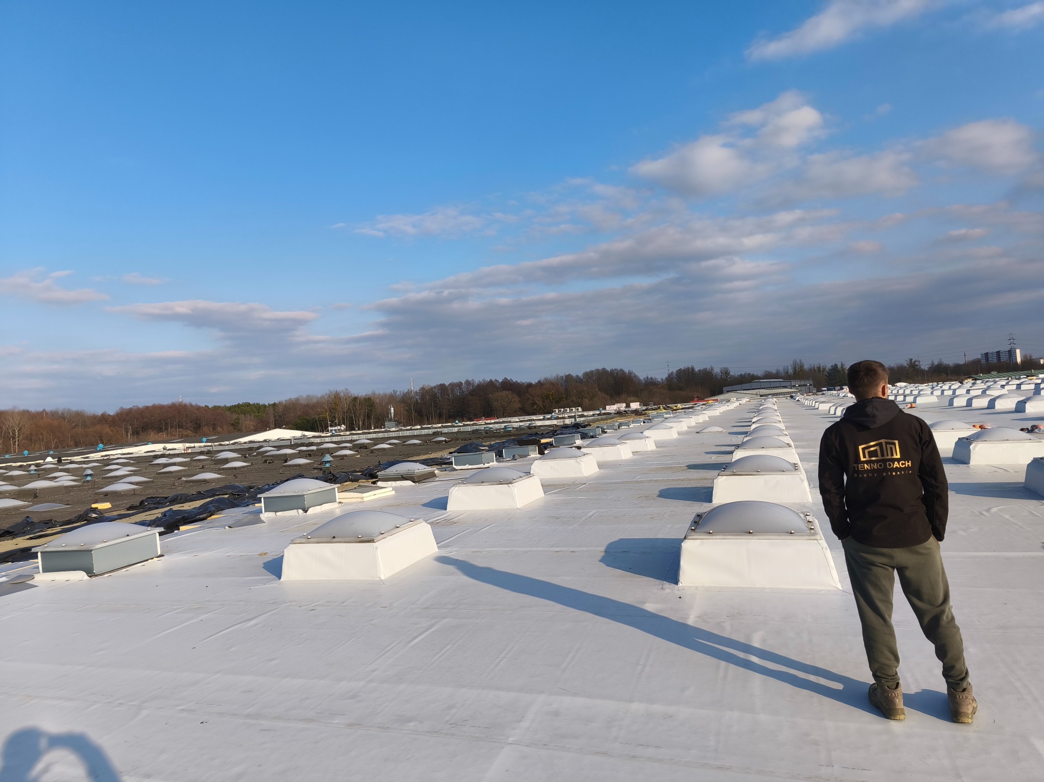 Biały dach z licznymi świetlikami, z sylwetką dekarza w tle, w słoneczny dzień. Widok z góry na rozległą powierzchnię dachu. Logo firmy na bluzie pracownika.