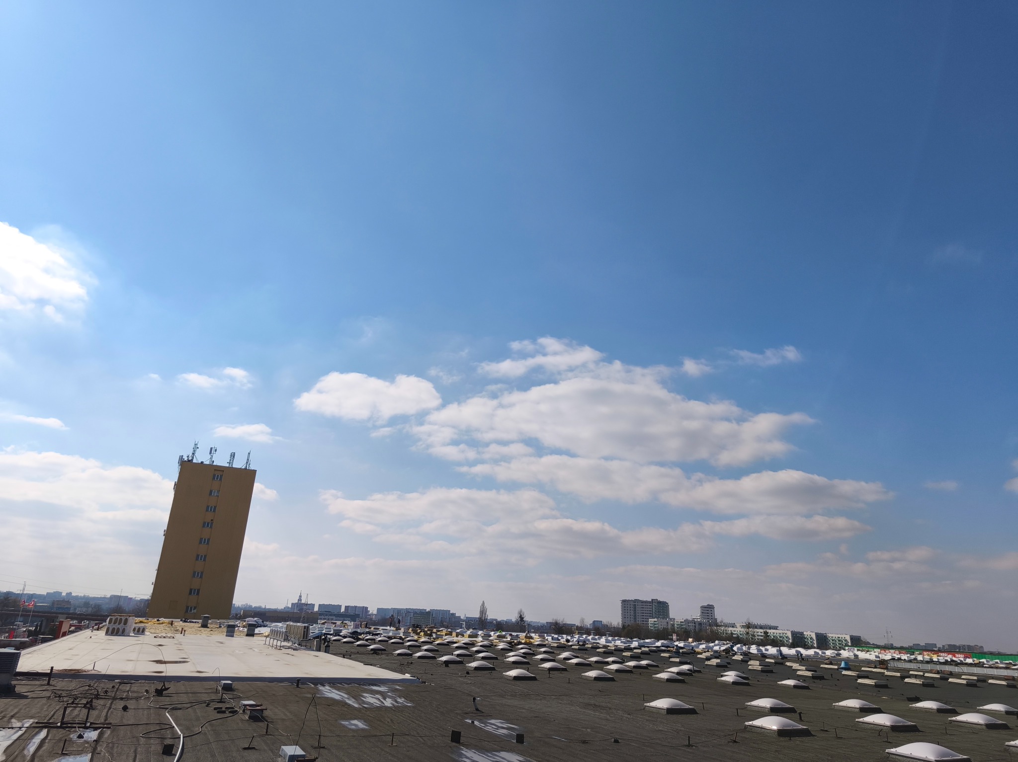 Widok z dachu pokrytego papą z licznymi świetlikami kopułowymi, w tle budynki miejskie i wieża telekomunikacyjna pod błękitnym niebem z chmurami.