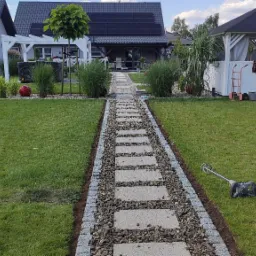 Ogród z ścieżką z płyt betonowych obramowaną kamieniami, prowadzącą do domu z panelami słonecznymi na dachu, w tle biała altana i pracownik z narzędziami.