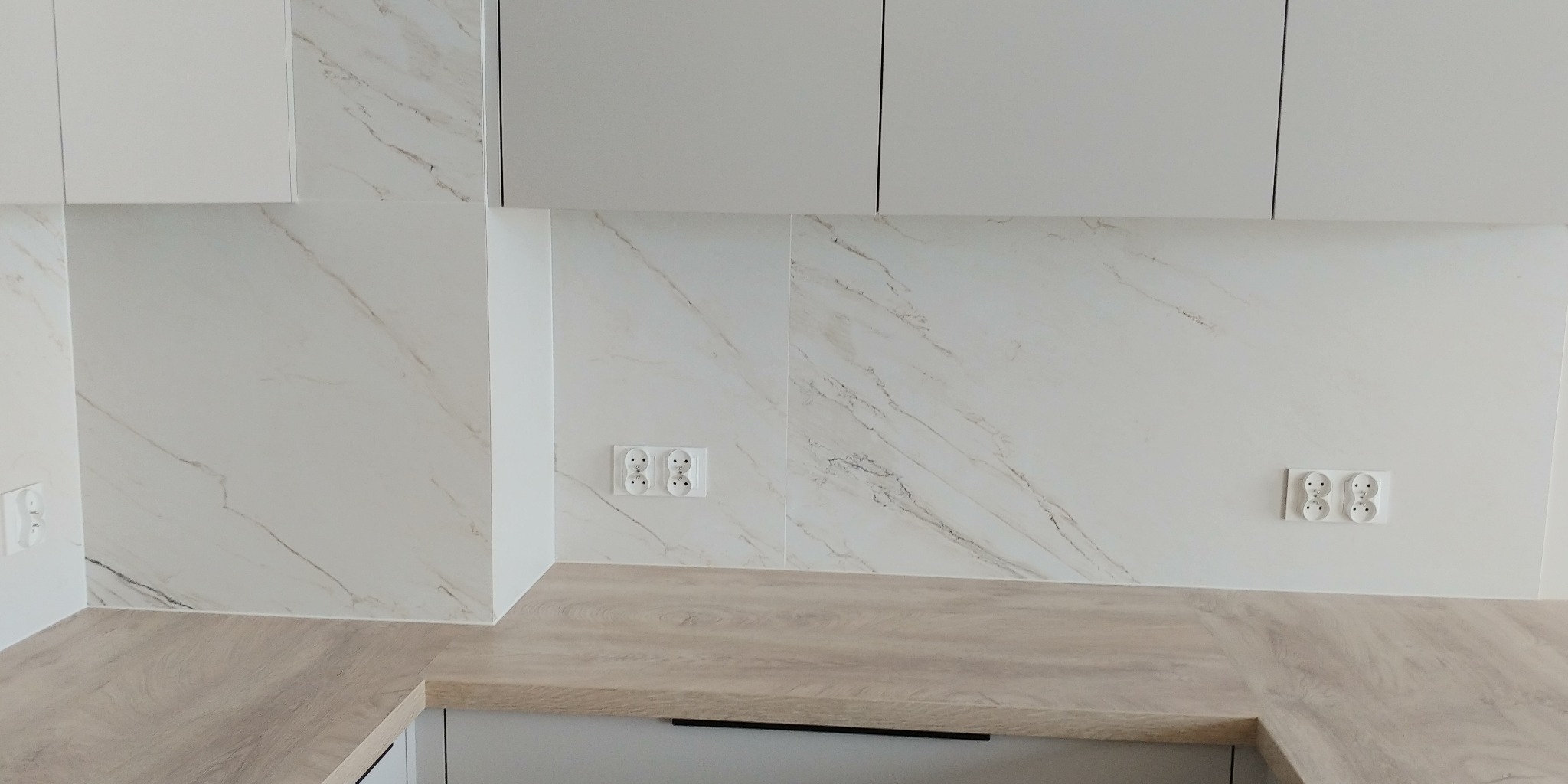 Białe płytki imitujące marmur na ścianie nad blatem kuchennym, dwa podwójne gniazdka elektryczne, minimalistyczne białe szafki wiszące.