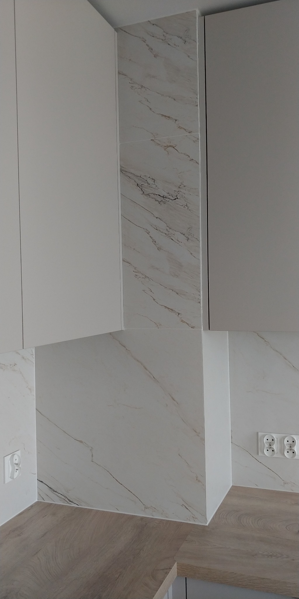 Fragment kuchni po remoncie: białe szafki z blatem imitującym drewno, ściana wykończona płytkami imitującymi marmur z widocznymi gniazdkami elektrycznymi.