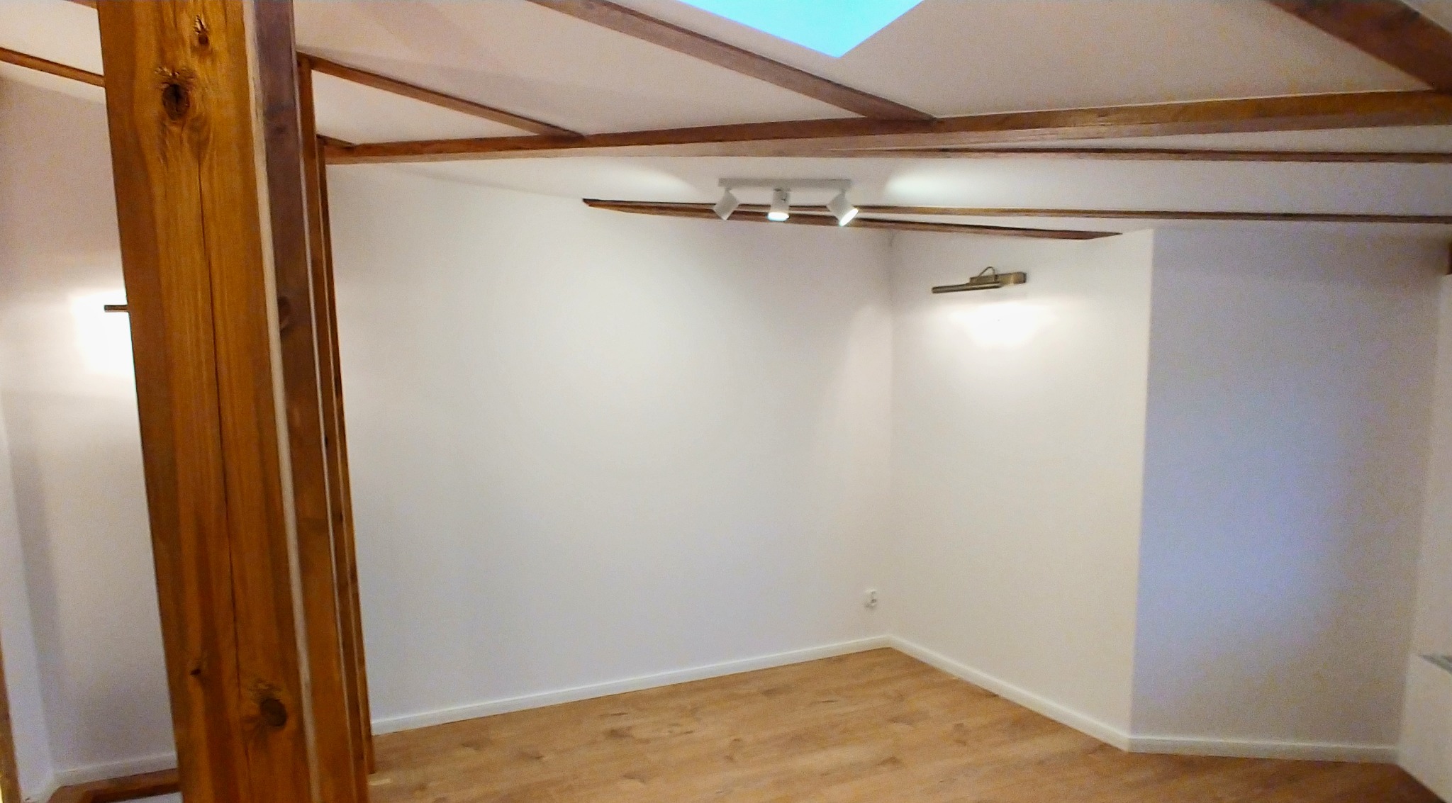 Pusty pokój z drewnianą podłogą i belek stropowych, oświetlony lampami sufitowymi i kinkietem, częściowo widoczna drewniana konstrukcja pionowa