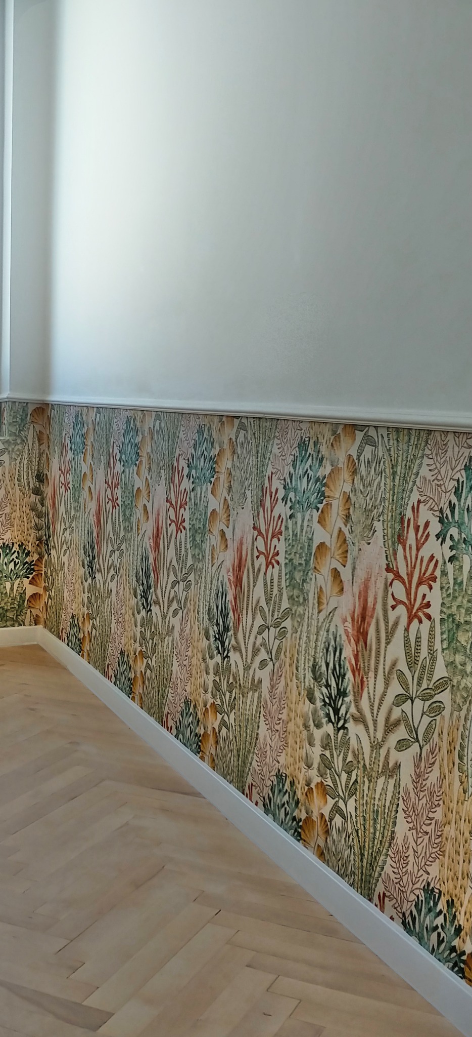 Ściana z wzorzystą tapetą w stylu botanicznym, świeżo położona w Gdańsku, z jasną listwą przypodłogową i parkietem ułożonym w jodełkę.