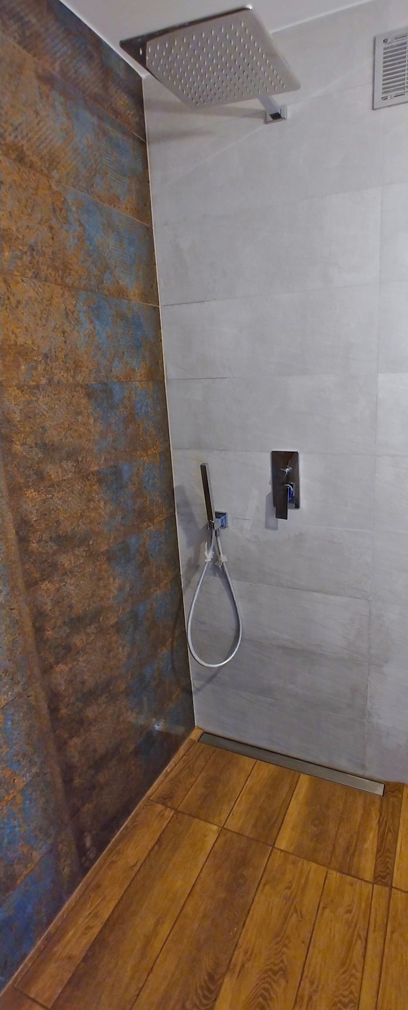 Nowoczesna kabina prysznicowa z deszczownicą, wykończona płytkami imitującymi rdzę na jednej ścianie i szarymi płytkami na pozostałych, z drewnopodobną podłogą i odpływem liniowym.