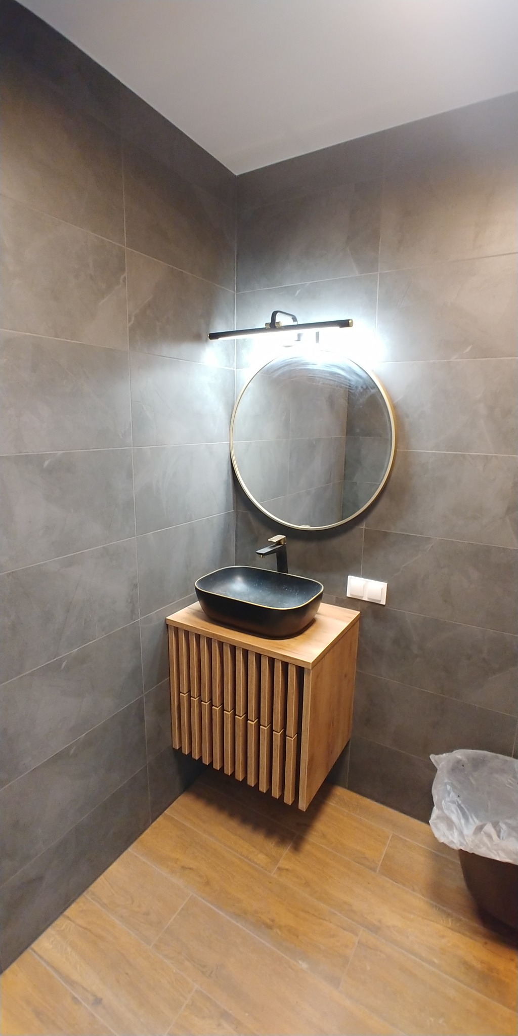 Nowoczesna łazienka z szarymi płytkami, drewnianą szafką pod umywalkę z lamelami, okrągłym lustrem z podświetleniem LED i czarną umywalką nablatową.