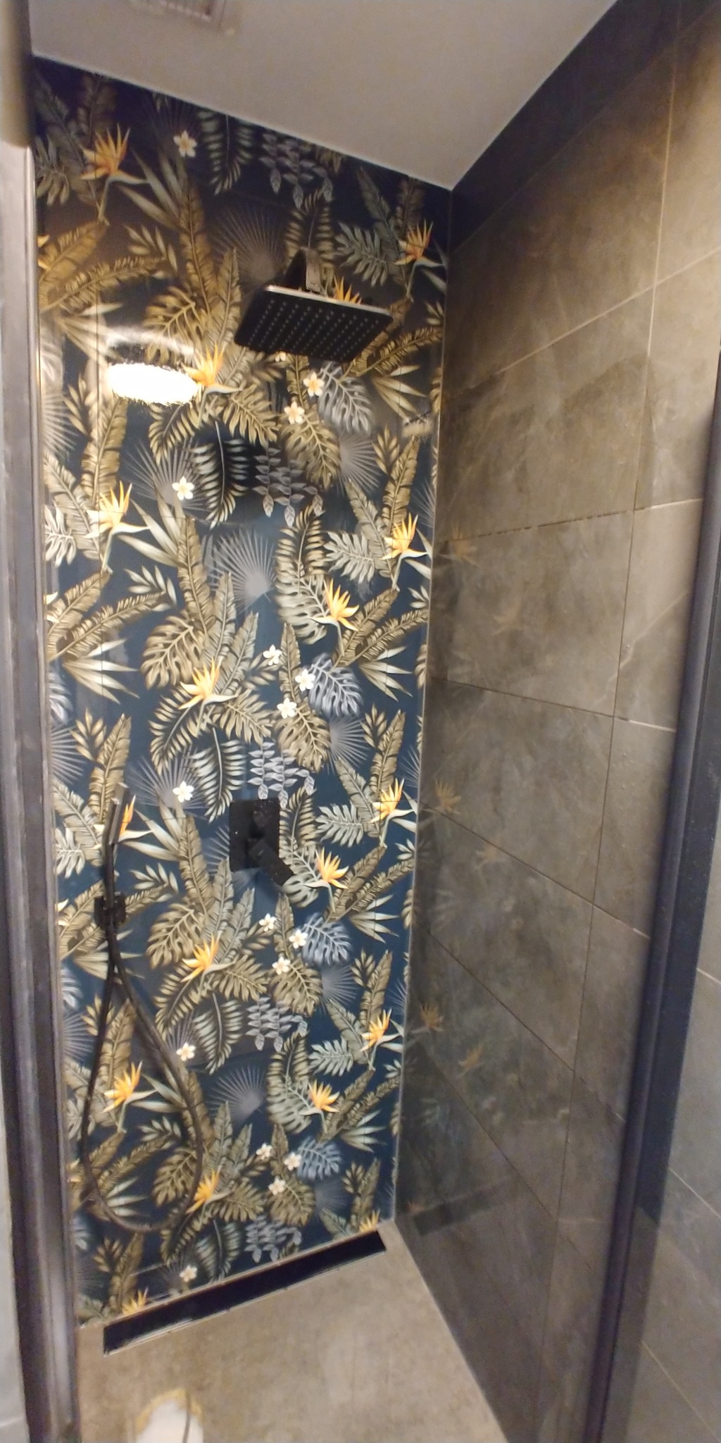 Kabina prysznicowa z egzotycznym motywem liści i kwiatów na ścianie, czarna deszczownica i bateria, szare płytki na sąsiedniej ścianie.