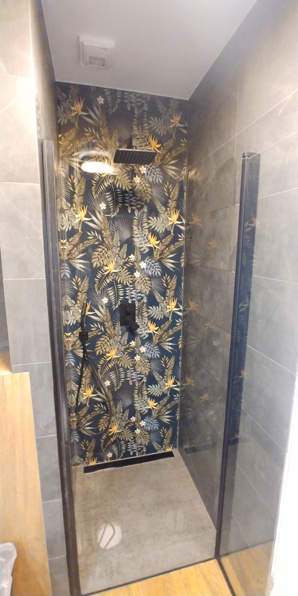 Nowoczesna kabina prysznicowa z deszczownicą, ściana w tropikalne liście, szare płytki na bocznych ścianach, szklane drzwi kabiny.