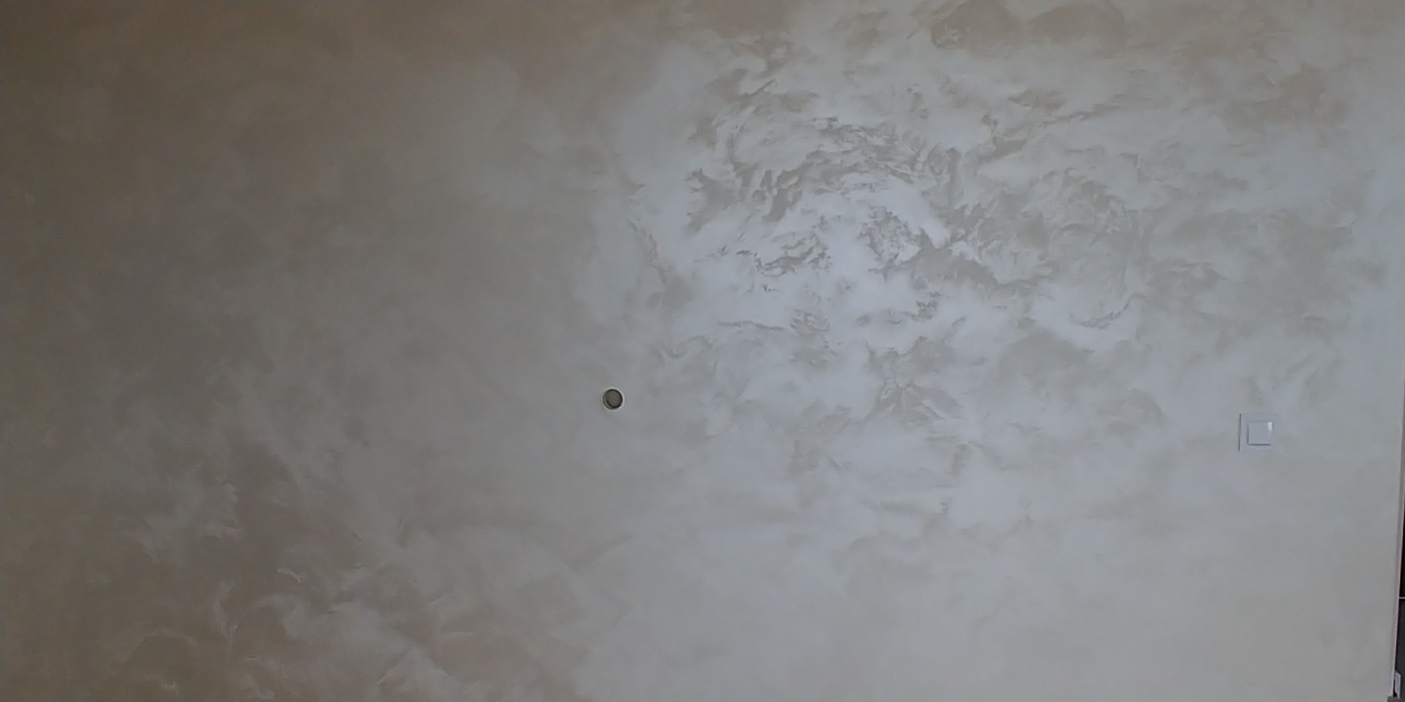 Ściana z dekoracyjną, beżowo-szarą farbą imitującą marmurkowy wzór, widoczny otwór na gniazdko i włącznik światła.