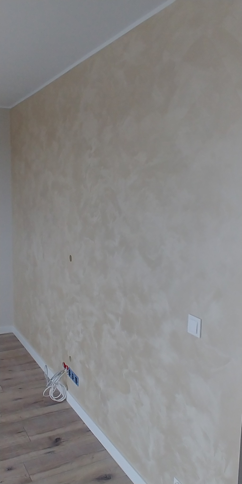 Ściana w pokoju po malowaniu dekoracyjnym z efektem perłowym, widoczne gniazdka elektryczne i fragment podłogi z paneli.