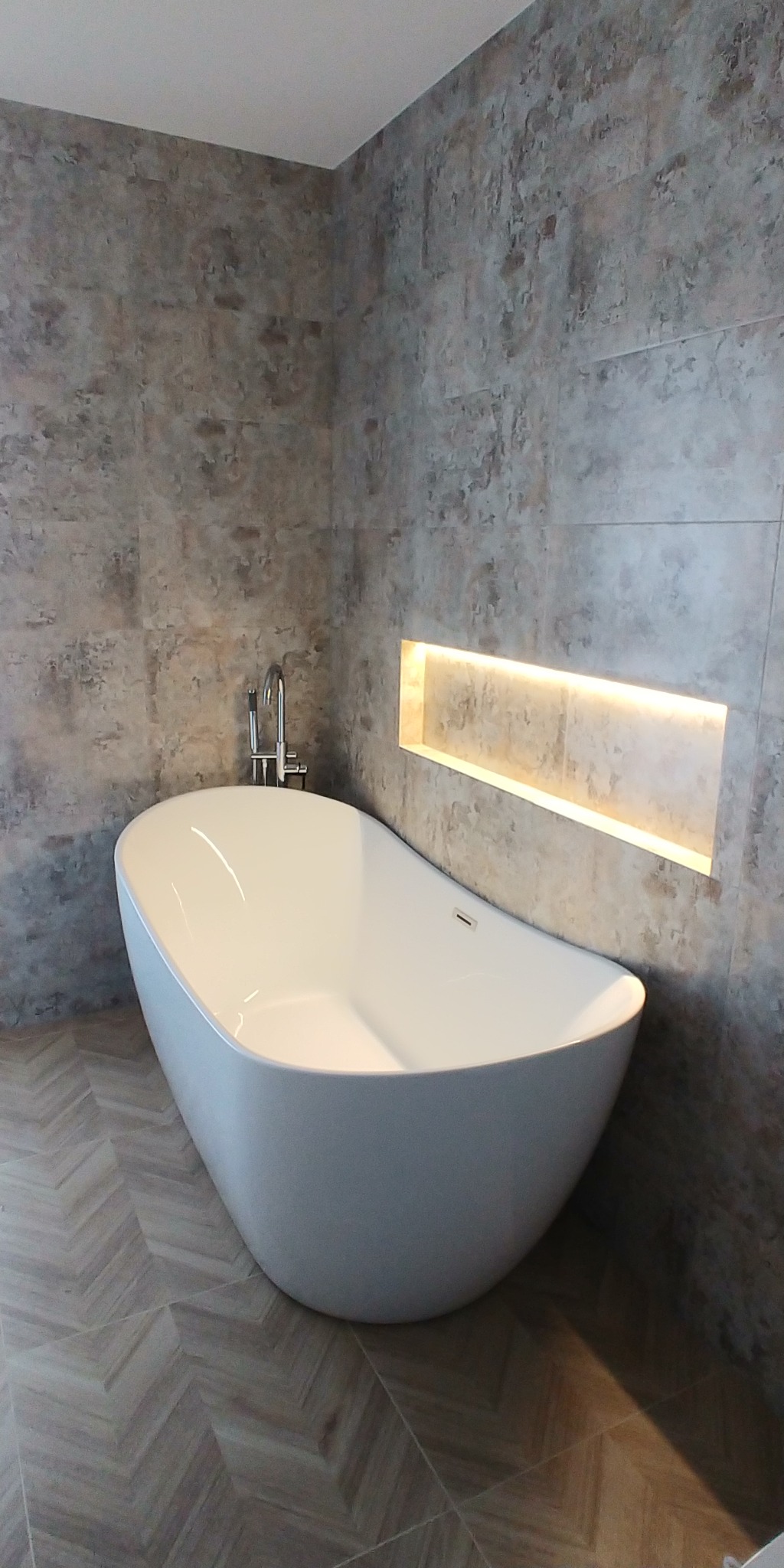 Wolnostojąca wanna z chromowaną armaturą na tle ściany z szarych płytek imitujących kamień, z podświetloną wnęką, podłoga wyłożona parkietem w jodełkę.