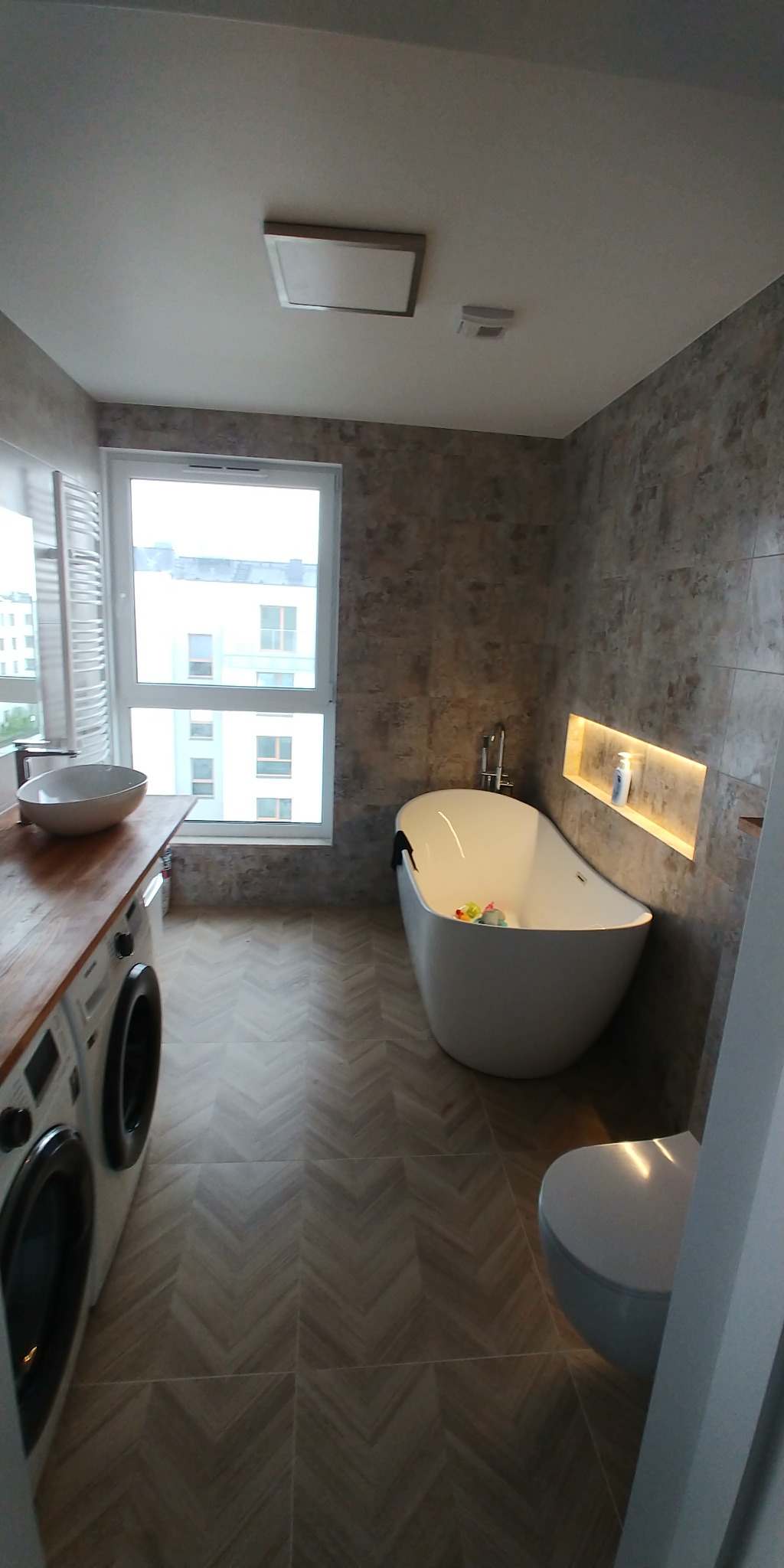 Nowoczesna łazienka z wanną wolnostojącą, pralką pod blatem z umywalką nablatową i toaletą, wykończona płytkami w odcieniach szarości ułożonymi w jodełkę na podłodze i imitującymi kamień...