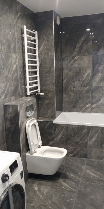 Nowoczesna łazienka z szarymi płytkami imitującymi marmur, podwieszana toaleta z otwartą klapą, biały grzejnik drabinkowy i pralka w rogu.