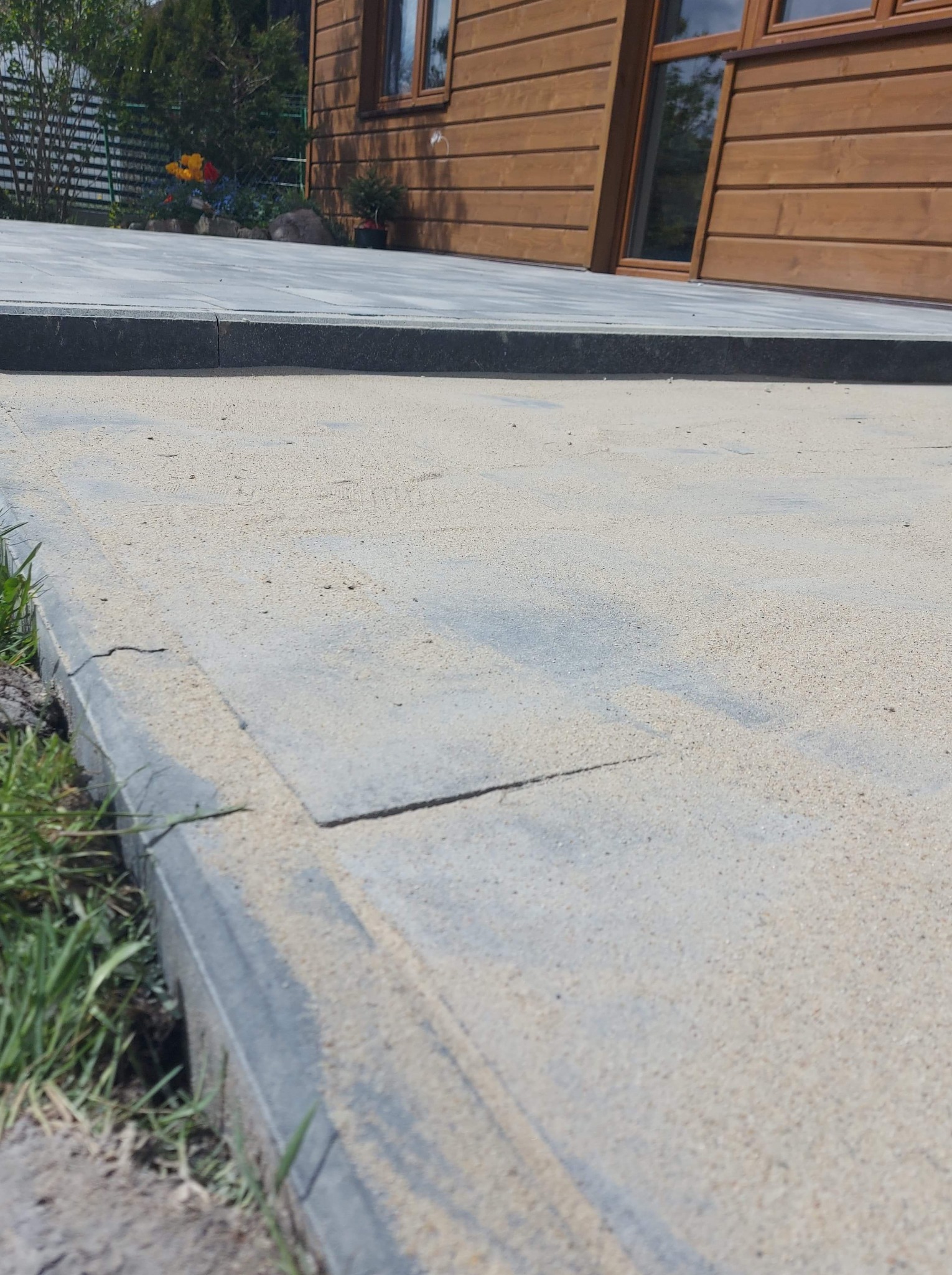 Szara kostka brukowa ułożona przy drewnianym domu, z widocznym piaskiem wypełniającym szczeliny i niskim obrzeżem trawnika.