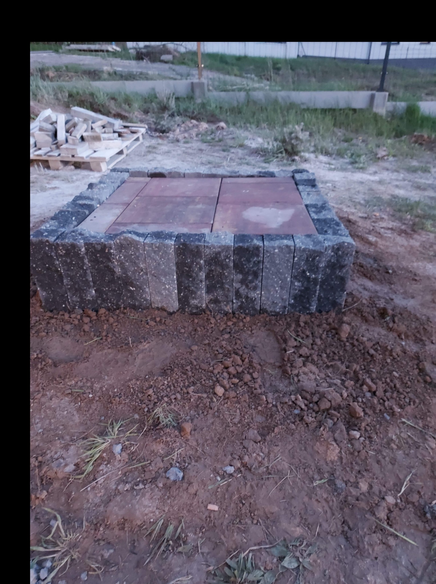 Niewielki placyk z kostki brukowej w trakcie budowy, otoczony obrzeżem z ciemnych kostek, widoczne nierówne podłoże z ziemi i trawy, w tle paleta z kostką.
