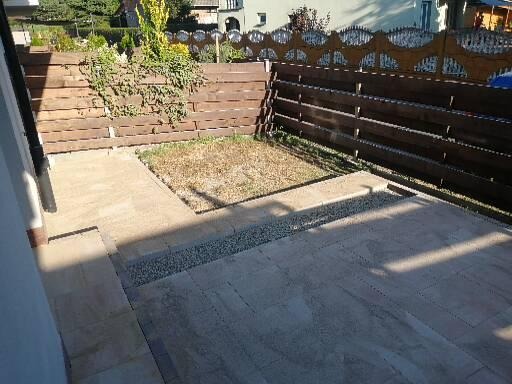 Wyłożony jasnymi płytami taras z drenażem żwirowym przy drewnianym ogrodzeniu z dekoracyjnym wykończeniem, przylegający do trawnika.