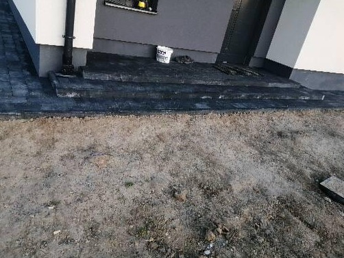 Ułożone, ciemne stopnie z kostki prowadzące do drzwi wejściowych domu, widoczny fragment chodnika z kostki i przygotowany teren pod dalsze prace.