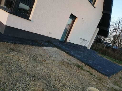 Ukończona ścieżka z ciemnej kostki brukowej prowadząca do domu z białymi ścianami, widoczna część podwórka z drobnym żwirem.