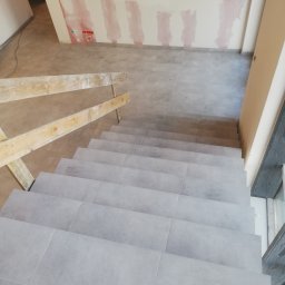 Kazimierz Maciak - Schody wyłożone szarymi płytkami, widok z góry, z fragmentem drewnianej balustrady i ścianą z widocznymi śladami po gładzi.