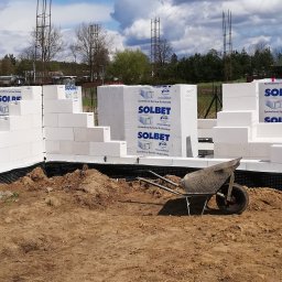 Kazimierz Maciak - Budowa domu w trakcie wznoszenia ścian z białych bloczków Solbet, widoczna taczka z piaskiem i wystające pręty zbrojeniowe.