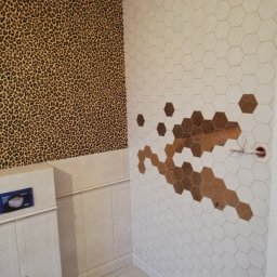 Kazimierz Maciak - Ściana łazienki z częściowo ułożonymi białymi, heksagonalnymi płytkami z wstawkami brązowych płytek tworzących wzór, sąsiednia ściana pokryta tapetą we wzór panterki, widoczna zabudowa podtynkowego...