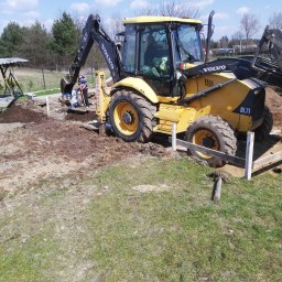 Kazimierz Maciak - Żółta koparko-ładowarka Volvo BL71 w trakcie prac ziemnych przy fundamentach, widoczni dwaj pracownicy z łopatami, drewniane szalunki, ziemia i trawa, błękitne niebo z chmurami w tle.