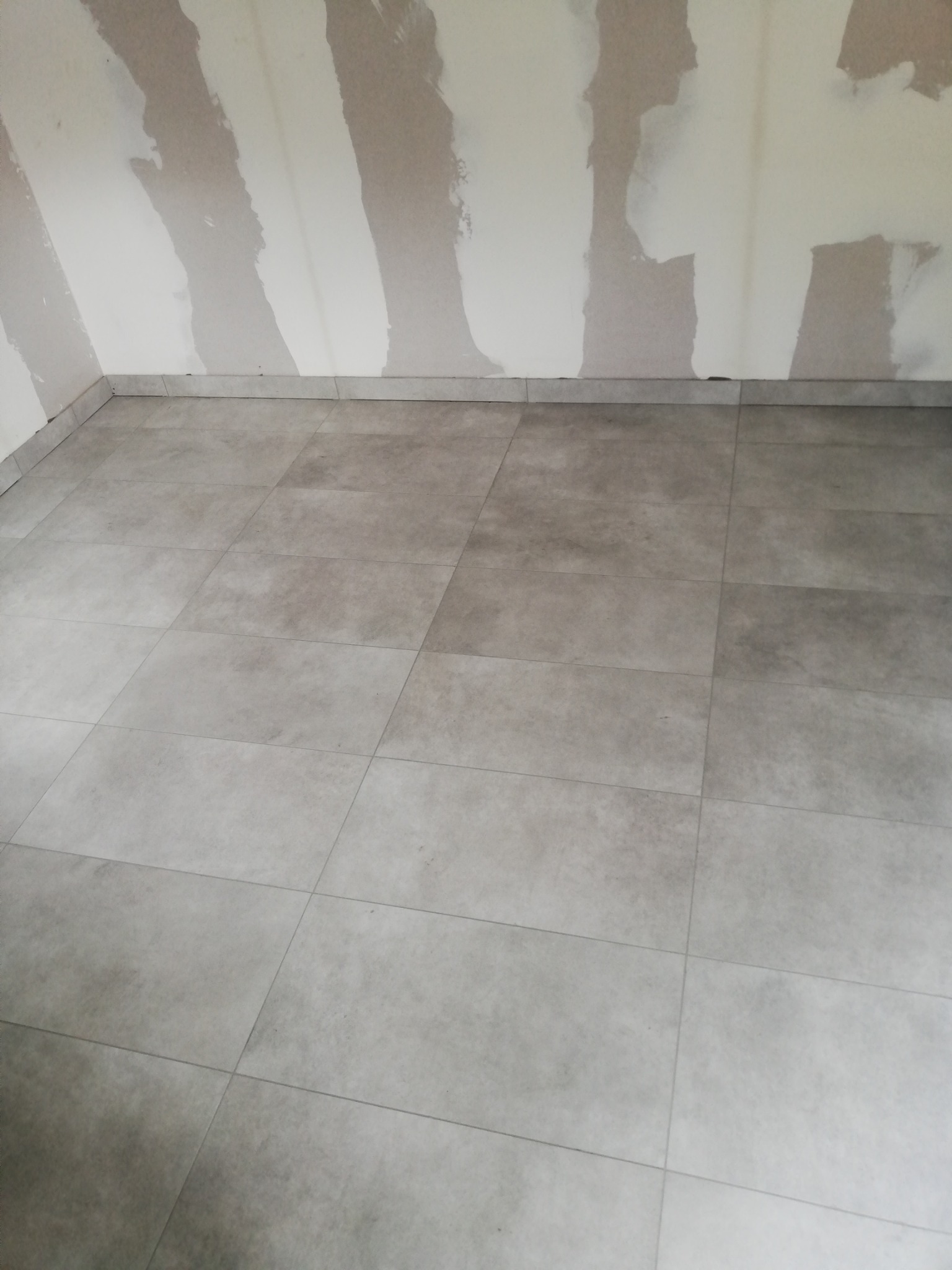 Wykończona podłoga z szarych, kwadratowych płytek w pomieszczeniu w trakcie remontu, ściany z widocznymi śladami szpachlowania.