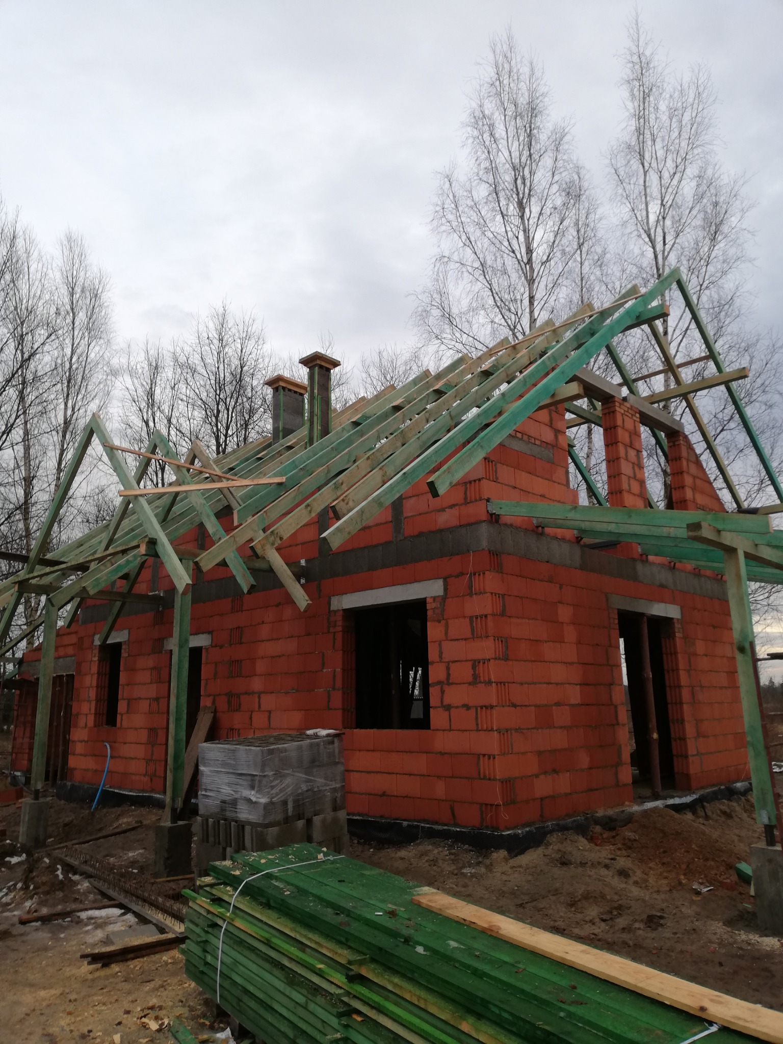 Surowy budynek z czerwonej cegły w trakcie budowy, z widoczną konstrukcją dachu z impregnowanego drewna, na tle bezlistnych drzew i pochmurnego nieba.