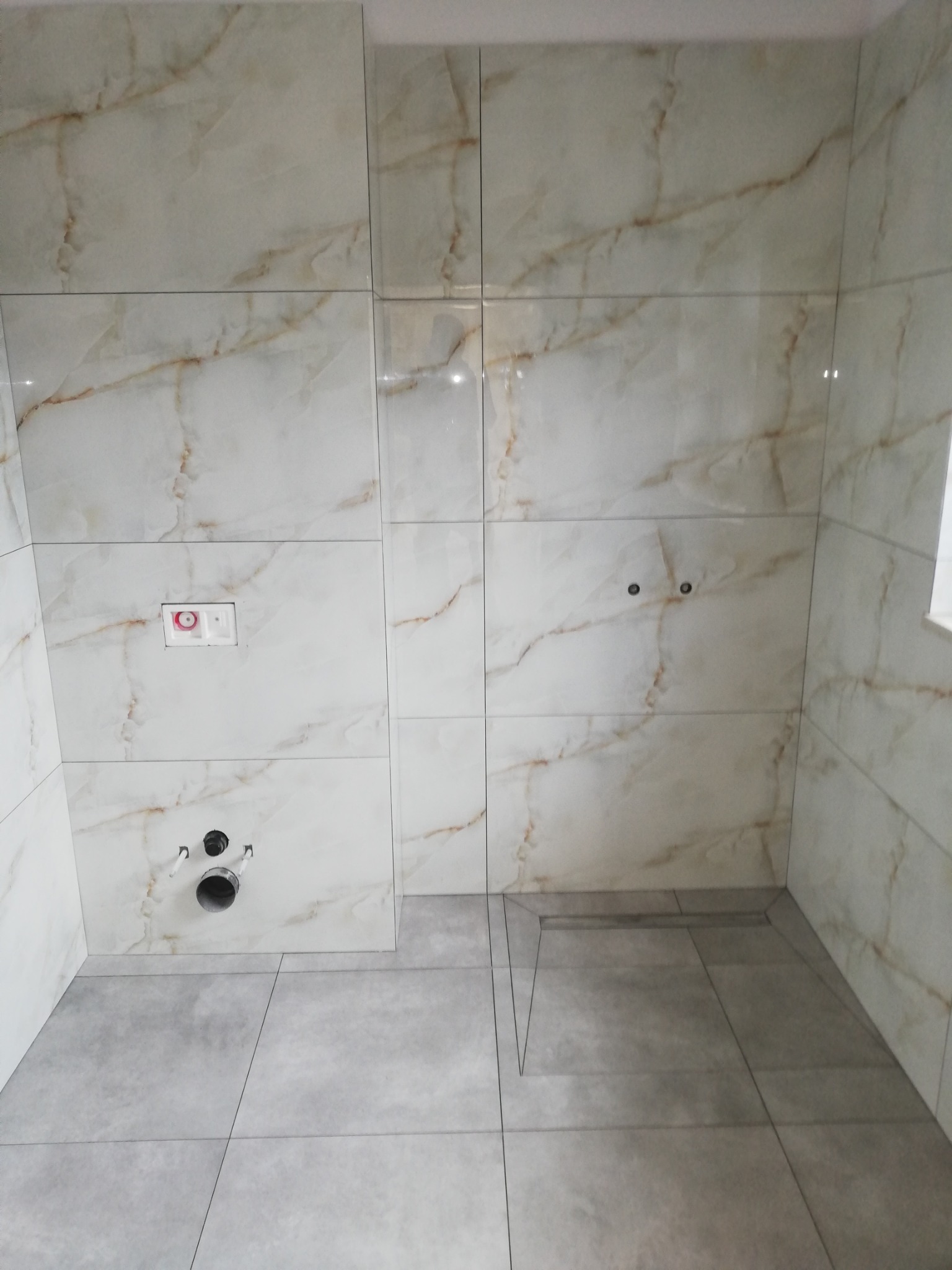 Wykończona łazienka z beżowo-złotymi płytkami imitującymi marmur na ścianach i szarymi płytkami podłogowymi, widoczne przygotowane przyłącza wodno-kanalizacyjne.