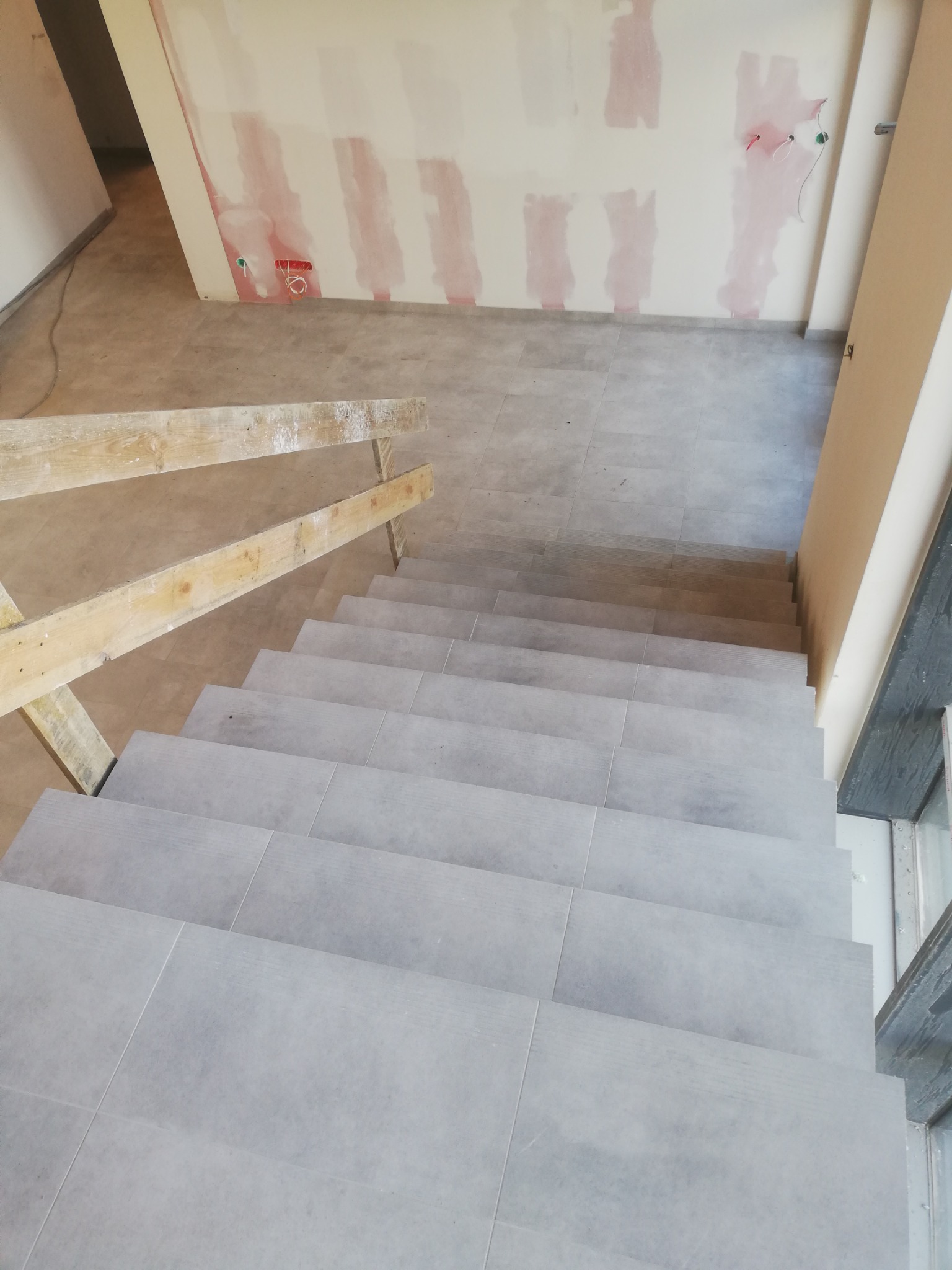 Schody wyłożone szarymi płytkami, widok z góry, z fragmentem drewnianej balustrady i ścianą z widocznymi śladami po gładzi.