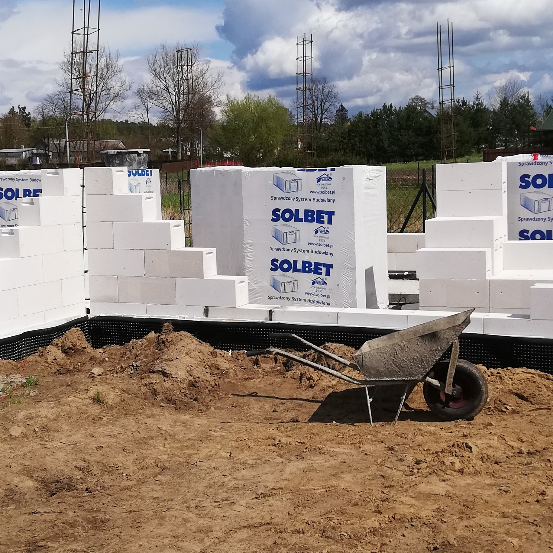 Budowa domu w trakcie wznoszenia ścian z białych bloczków Solbet, widoczna taczka z piaskiem i wystające pręty zbrojeniowe.
