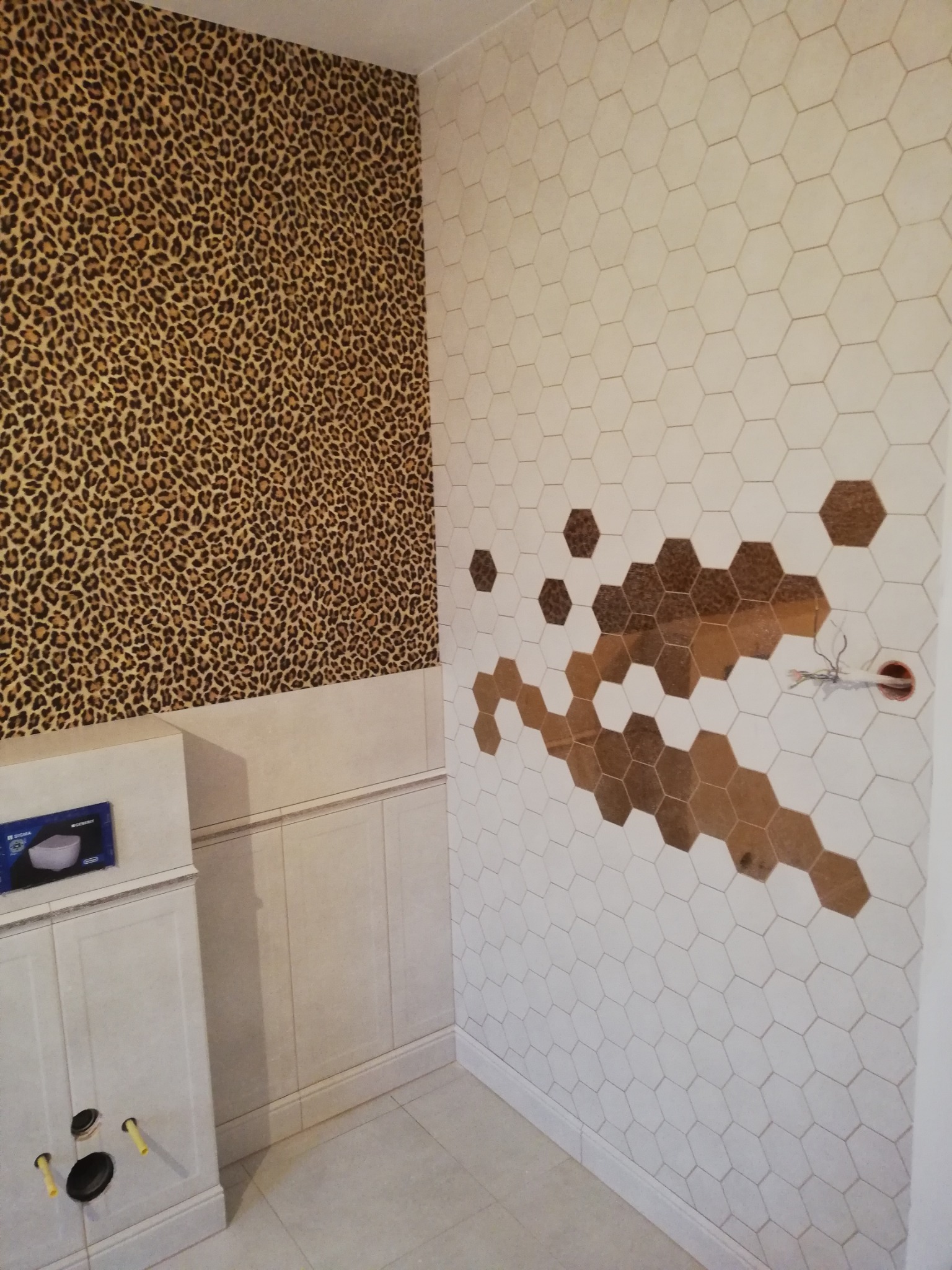Ściana łazienki z częściowo ułożonymi białymi, heksagonalnymi płytkami z wstawkami brązowych płytek tworzących wzór, sąsiednia ściana pokryta tapetą we wzór panterki, widoczna zabudowa podtynkowego...