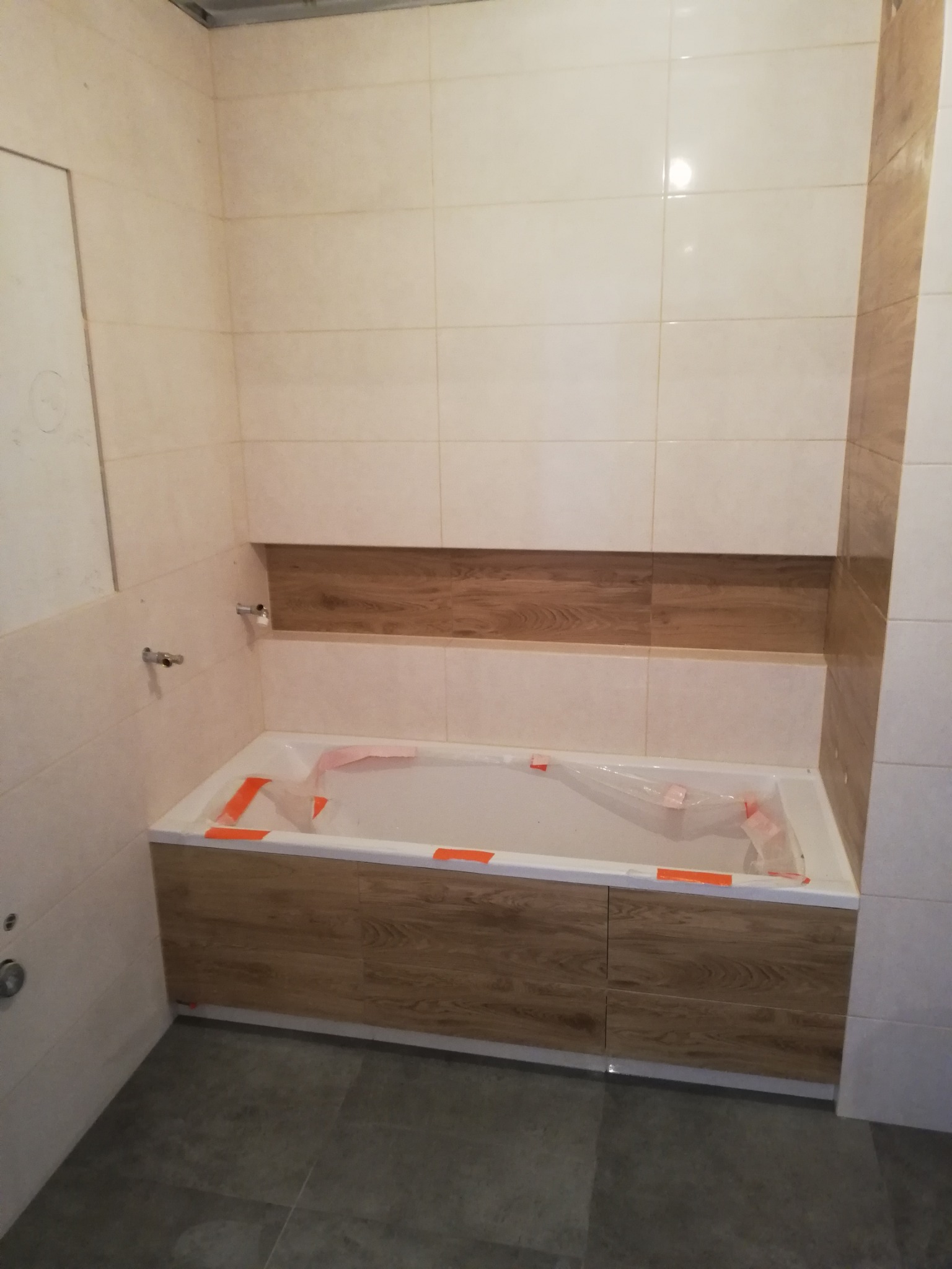 Wykończona łazienka z wanną obudowaną panelami imitującymi drewno, jasne płytki na ścianach z dekoracyjnym pasem w podobnym stylu, szare płytki na podłodze, widoczne zabezpieczenia w wannie.