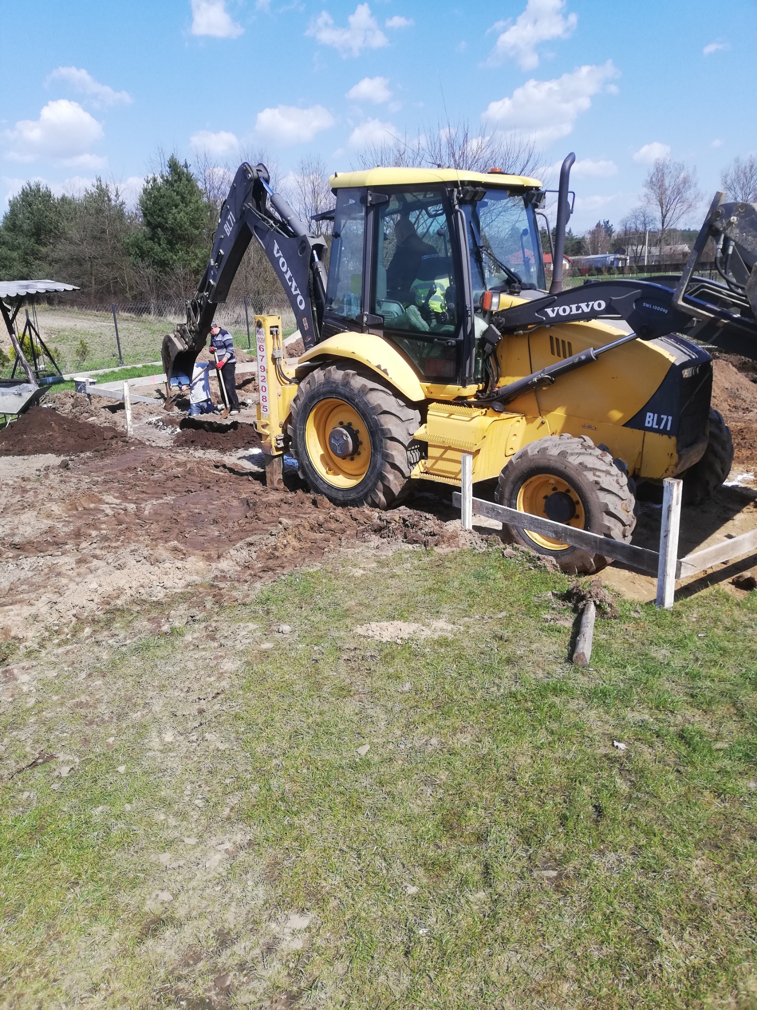 Żółta koparko-ładowarka Volvo BL71 w trakcie prac ziemnych przy fundamentach, widoczni dwaj pracownicy z łopatami, drewniane szalunki, ziemia i trawa, błękitne niebo z chmurami w tle.