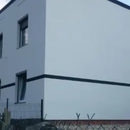 Ocieplony budynek z białymi ścianami i czarnymi pasami dekoracyjnymi, widoczny przez metalowe ogrodzenie i kamienny murek.