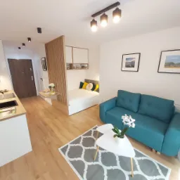 Aranżacja wnętrza małego mieszkania z aneksem kuchennym, sypialnią oddzieloną drewnianą ścianką, turkusową sofą i stolikiem kawowym na wzorzystym dywanie.