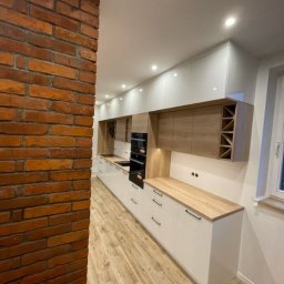 Plan na meble - Długa, biała kuchnia z drewnianymi blatami i górnymi szafkami, widoczna z perspektywy pod kątem, z ceglaną ścianą po lewej stronie i drewnianą podłogą.