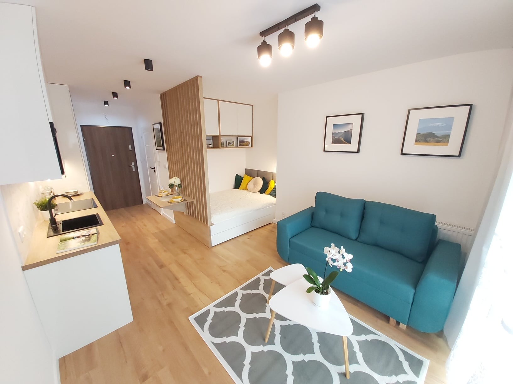 Aranżacja wnętrza małego mieszkania z aneksem kuchennym, sypialnią oddzieloną drewnianą ścianką, turkusową sofą i stolikiem kawowym na wzorzystym dywanie.