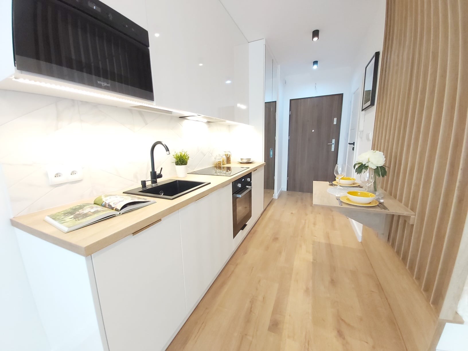 Wnętrze małej kuchni z białymi frontami, drewnianym blatem, czarną armaturą i wbudowanym sprzętem AGD, uzupełnione dekoracjami w postaci książki, rośliny w doniczce i zastawy stołowej w odcieniach...