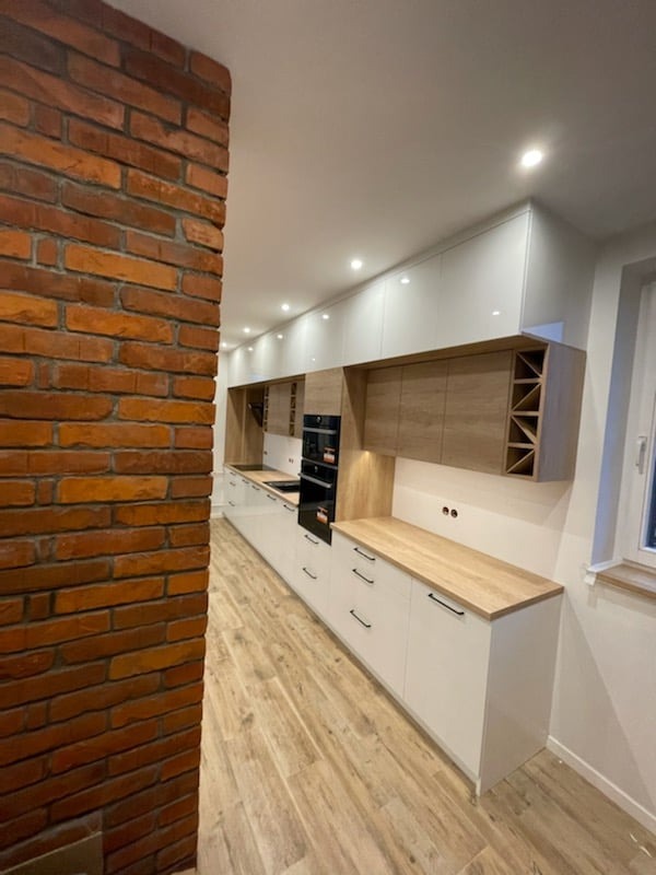 Długa, biała kuchnia z drewnianymi blatami i górnymi szafkami, widoczna z perspektywy pod kątem, z ceglaną ścianą po lewej stronie i drewnianą podłogą.