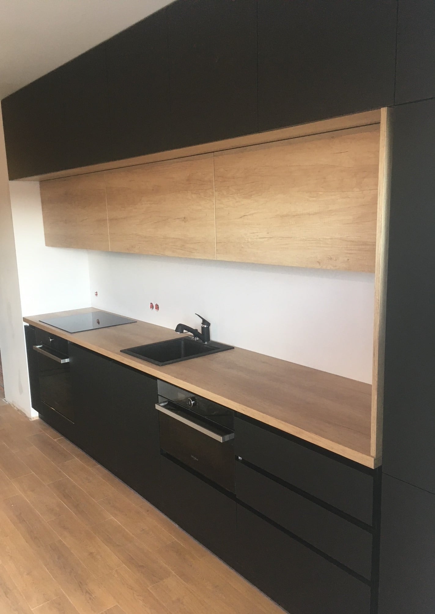 Nowoczesna kuchnia z czarnymi szafkami dolnymi i drewnopodobnymi szafkami górnymi, czarny zlew i płyta indukcyjna w blacie z drewnopodobnym wykończeniem, widoczne niedokończone wykończenia ściany.