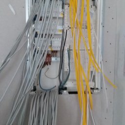 Jakub Warmijak - Rozdzielnica elektryczna w ścianie z dużą ilością przewodów w kolorach szarym i żółtym, widoczne elementy zabezpieczeń i złączki.