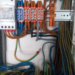 Jakub Warmijak - Rozdzielnica elektryczna z widocznymi przewodami różnego koloru, złączkami, bezpiecznikami i ogranicznikiem przepięć, na białym tle.