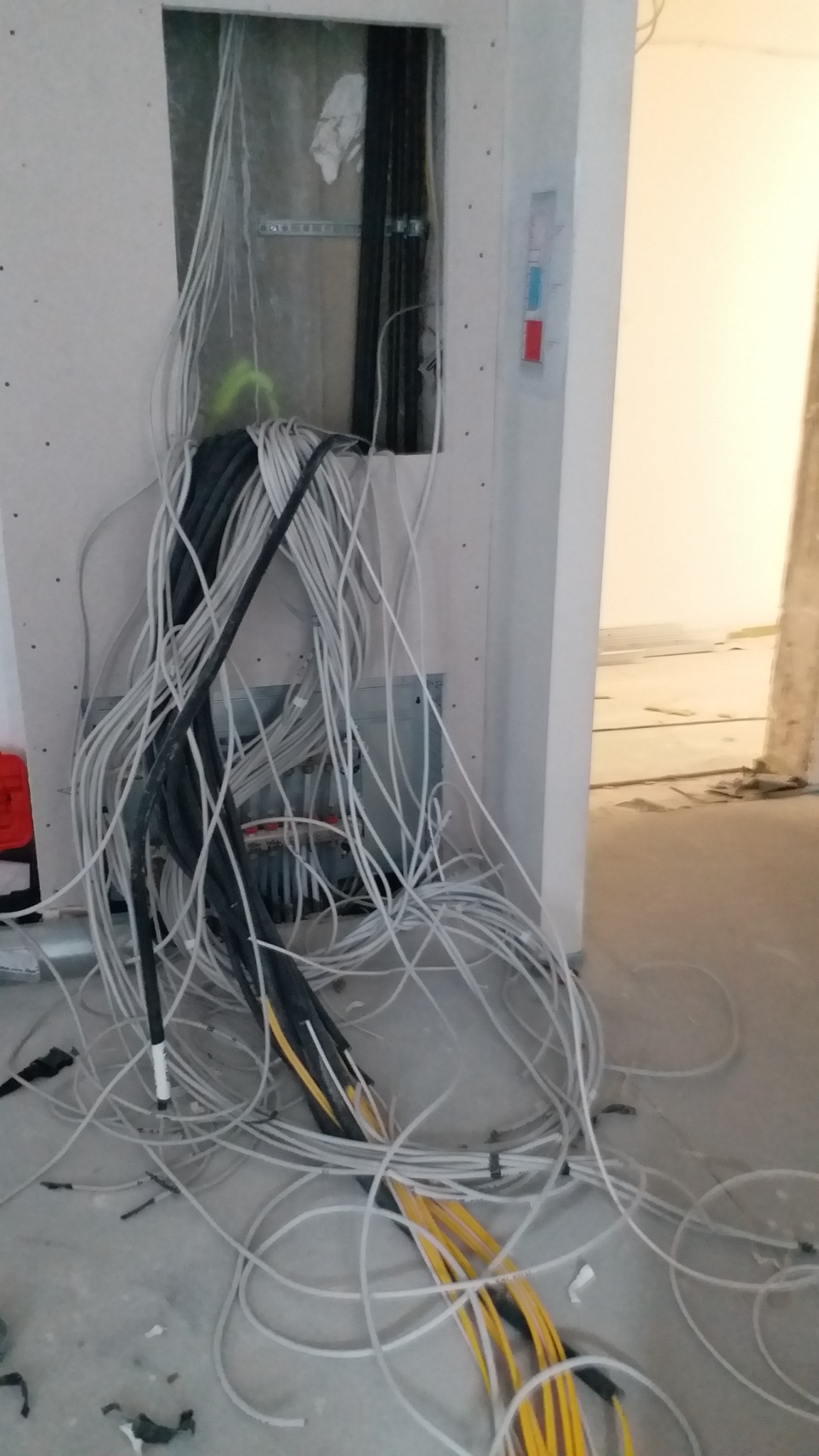 Wiązka przewodów elektrycznych różnych kolorów wychodząca z otworu w ścianie, widoczne elementy montażowe wewnątrz otworu, surowy stan wykończenia wnętrza.